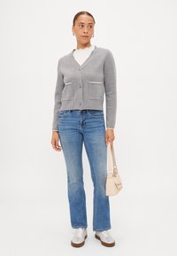 Γκρι πλεκτό πουλόβερ με V-λαιμόκοψη, δύο μπροστινές τσέπες και κρεμ λεπτομέρειες, συνδυασμένο με μπλε φαρδιά τζιν και μεταλλικά αθλητικά παπούτσια.