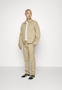Beige kort jakke kombinert med matchende bukser. Hvit t-skjorte under. Sorte joggesko med gule striper. Enfarget, tilfeldig design.