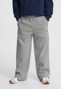 Graue Sweatpants mit lockerer Passform und elastischem Bund, versehen mit einem kleinen Logo an der Seite. Getragen mit weißen und grünen Sneakers.