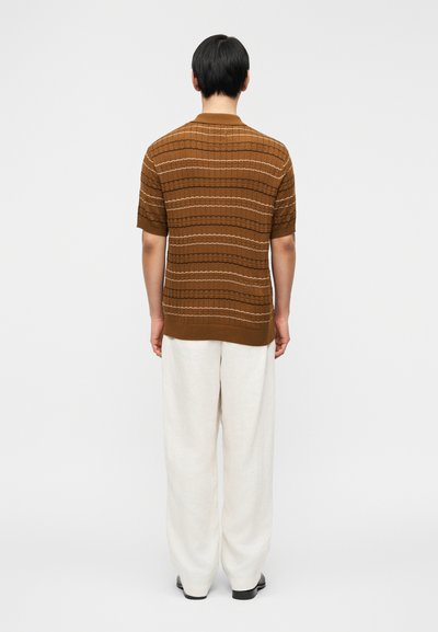 Άνδρας που στέκεται με την πλάτη προς τα εμάς, φοράει καφέ ριγέ πλεκτό κοντομάνικο πουκάμισο, λευκό άνετο παντελόνι και μαύρα παπούτσια σε απλό λευκό φόντο.