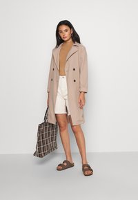 Lys beige dobbelknappet frakk, ribbet beige genser, hvite denimshorts, brune sandaler og en rutete totebag. Glatt tekstur.