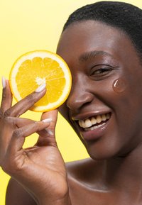 Uno sfondo giallo brillante presenta una persona sorridente che tiene una fetta d'arancia fresca e rotonda vicino al volto, mostrando una pelle sana e unghie lucide.