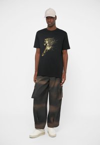 Black T-shirt with gold graphic, paired with dark cargo pants featuring a beige and brown pattern. Model wears a light beige cap and white shoes.