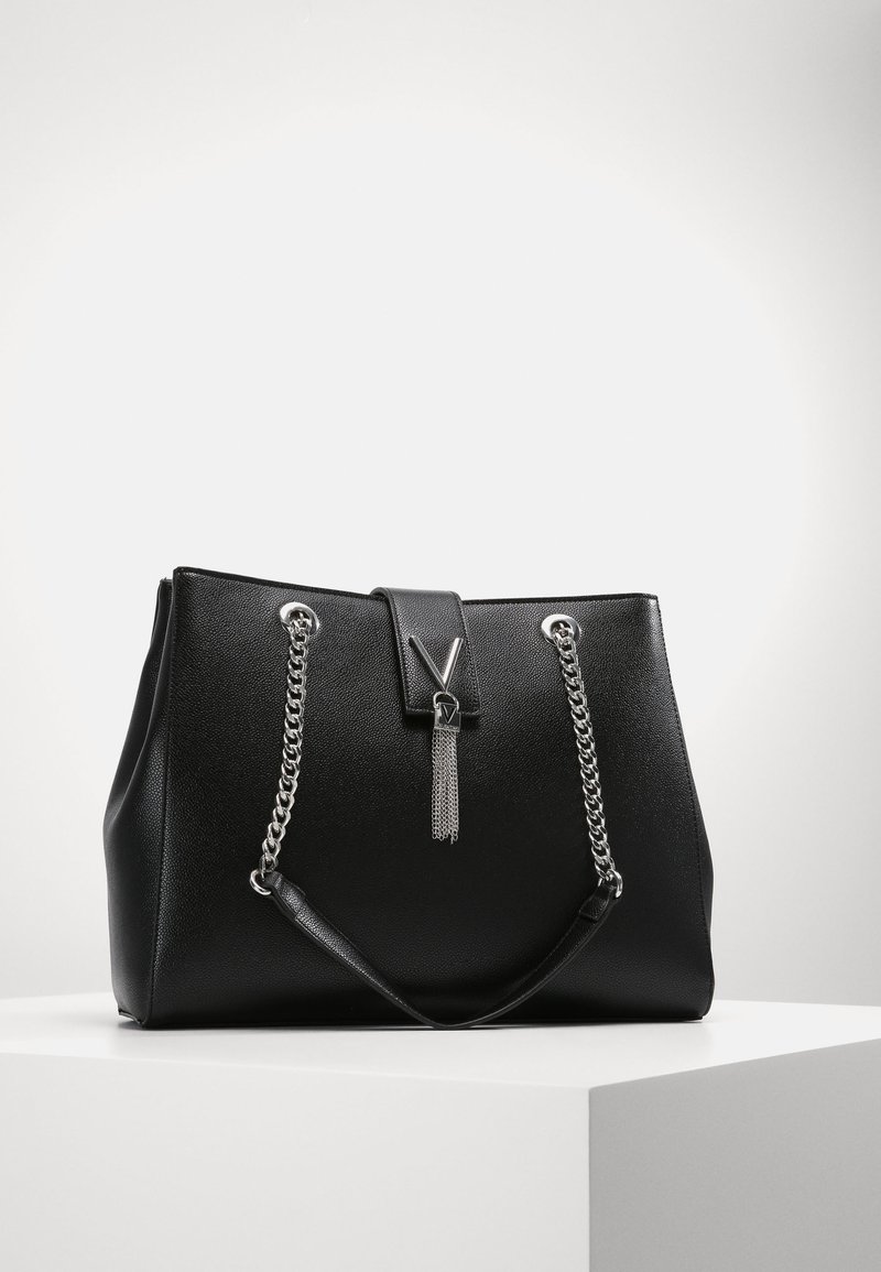 Sac fourre-tout en simili cuir noir avec des poignées en chaîne argentée, un rabat avant avec des accents métalliques et une surface texturée.