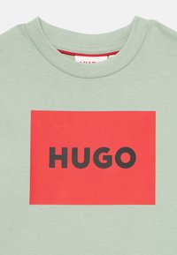 Zelené bavlnené tričko s veľkým červeným obdĺžnikovým grafickým motívom a čiernym slovom "HUGO" potlačeným v tučných, blokových písmenách.