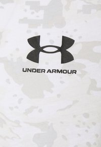 Camisa de manga corta en gris claro con un patrón de camuflaje. Presenta un logotipo de Under Armour en negro, exhibido de manera prominente en la parte frontal.