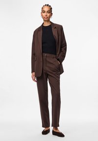 Brauner maßgeschneiderter Anzug mit einem einreihigen Blazer, geripptem schwarzen Oberteil und geraden Hosen. Akzentuiert mit dezenten Taschen und weichem Material.
