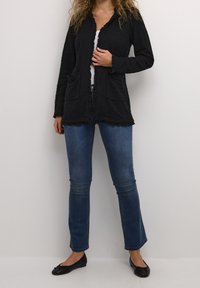 Czarny teksturowany kardigan z falbanami na krawędziach, kieszeniami z przodu i otwartym fasonem. Połączony z dopasowanymi niebieskimi dżinsami i czarnymi balerinami.