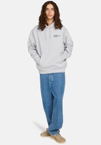 Sudadera gris con un bolsillo frontal, que presenta un pequeño logotipo azul. Se lleva con unos vaqueros holgados azules y zapatos marrón claro.