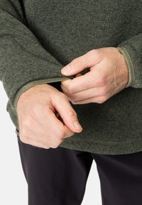 Cămașă lungă cu mânecă, texturată, de culoare verde închis, cu un tiv contrastant de verde mai deschis la manșete, parțial rulate, arătând mâinile care ajustează mâneca.