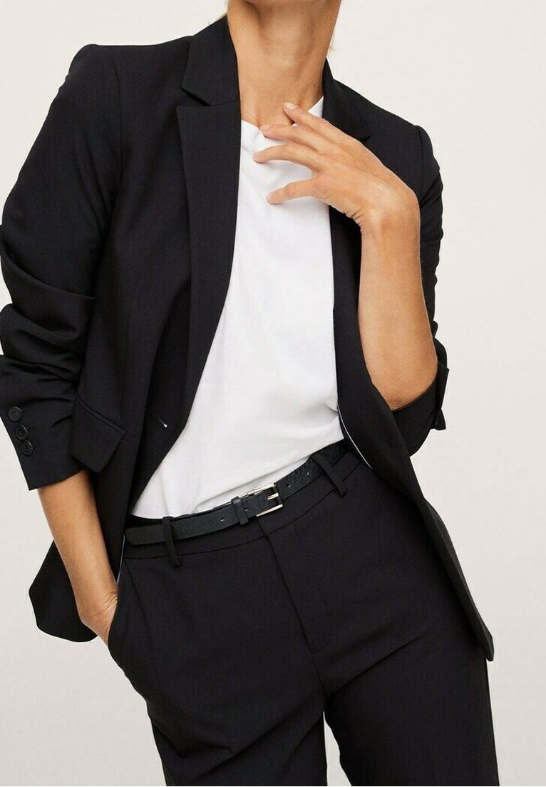 Femme portant un blazer noir aux manches retroussées, un pantalon noir, une ceinture noire et une chemise blanche, une main dans la poche, l'autre près du cou.