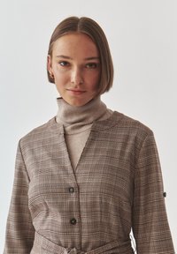 Abito a maniche lunghe con stampa a quadri in marrone e beige, caratterizzato da un profondo scollo a V e una vita annodata, indossato sopra un dolcevita beige.