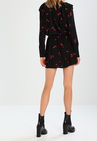 Robe noire à motifs floraux rouges, détails à volants sur les épaules, taille élastiquée, et associée à des bottines à talons noires.