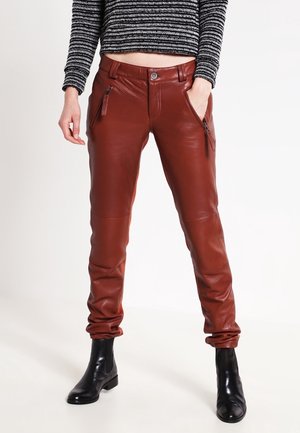 Vrouw draagt roestkleurige leren broek met ritszakken en zwarte enkellaarzen, gecombineerd met een zwart-wit gestreepte cropped trui.