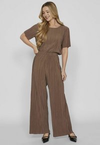 Completo marrone a due pezzi con pieghe, composto da una maglietta a maniche corte e pantaloni a gamba larga, realizzato in tessuto leggero. Scarpe nere con punta appuntita.
