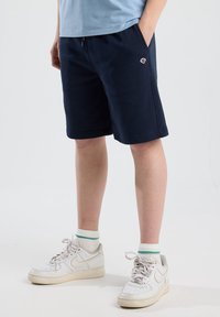 Laivansinis lygašortai su šoninėmis kišenėmis, elastinga juosta ir maža logotipo etikete. Sujungti su baltais sportbačiais ir žaliai dryžuotomis kojinėmis.