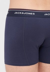 Mornarice plave bokserice napravljene od mekog materijala, s kontrastnim pojasom na kojem je bijeli logo "JACK & JONES", uokviren crvenim prugama.