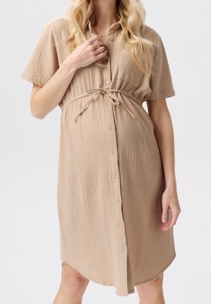 Femme portant une robe beige texturée boutonnée, à manches courtes et ceinture nouée à la taille, debout devant un fond uni.