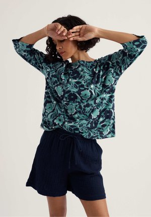 Vrouw met kort krullend haar, gekleed in een blauwgroene top met lange mouwen en bloemenprint en een marineblauwe, gestructureerde korte broek, poserend met opgetilde armen vlakbij haar gezicht.