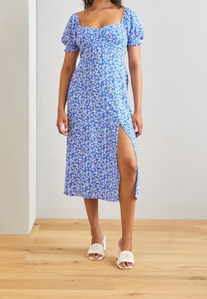 Kvinde iført en blå blomstret midi-kjole med pufærmer og et højtslids ved låret, kombineret med hvide flettede hælsandaler.