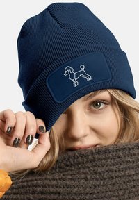 Navy knit beanie with textured pattern and rectangular patch featuring a white outline of a poodle. The fabric appears stretchy and warm.