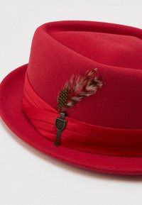 Piros filcből készült fedora, amelyhez illeszkedő piros szalagdísz tartozik, két barna és fekete foltos tollal díszítve, valamint egy fém kitűzővel az oldalon.