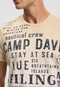 Beige cotton t-shirt featuring bold navy text, including “CAMP DAVID” and nautical-themed graphics. Soft texture with a crew neck design.