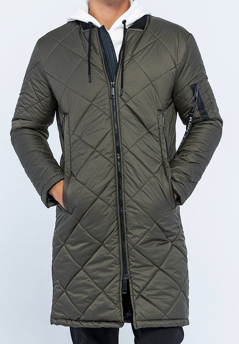 Lang, olivengrøn quiltet frakke med lynlås foran, hætte og lommer. Har et diamantmønster og snørre til justering.