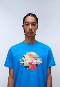 Modré bavlněné tričko s barevným kruhovým potiskem s abstraktními tvary a textem "Napapijri geographic" v bílé barvě přes hrudník.