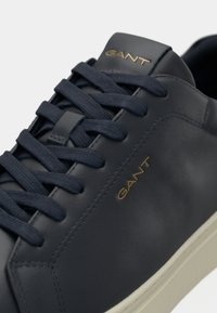 Adidași din piele bleumarin, cu șireturi tonale, un logo auriu pe lateral, textură netedă și o talpă din cauciuc de culoare deschisă în contrast.