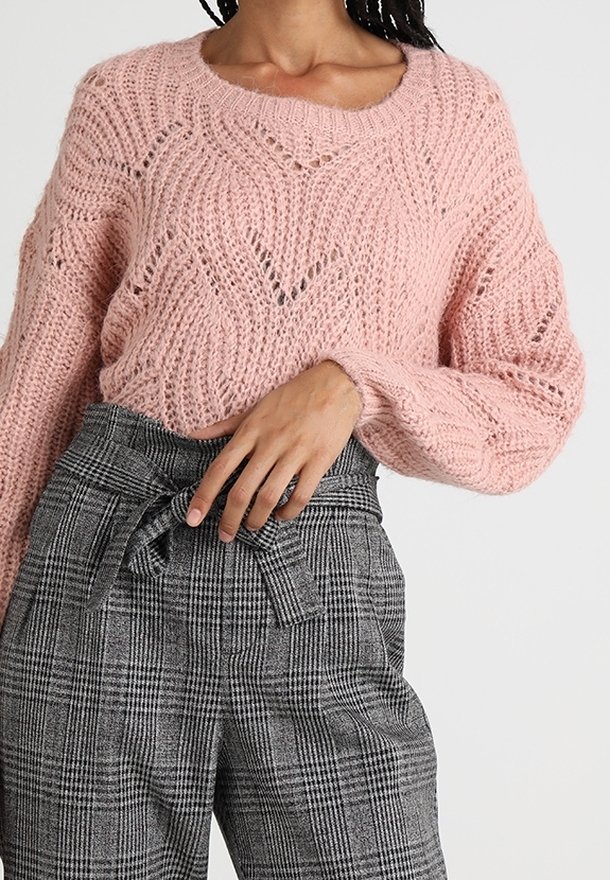 Kobieta ubrana w różowy sweter z teksturowanym splotem oraz wysokie szare spodnie w kratę z wiązanym paskiem z materiału.