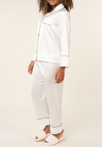 Ensemble de pyjama en satin blanc avec des détails en pipings noirs, comprenant un haut à manches longues avec des boutons et un pantalon droit, accompagné de chaussons blancs moelleux.