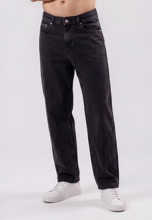 Mand iført falmede sorte jeans med lige ben og hvide sneakers står mod en ensfarvet baggrund.
