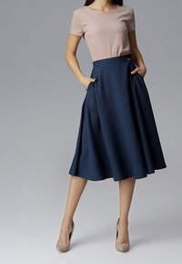 Femme portant un top beige à manches courtes, rentré dans une jupe midi bleu marine taille haute avec des poches, associée à des chaussures à talons beiges.