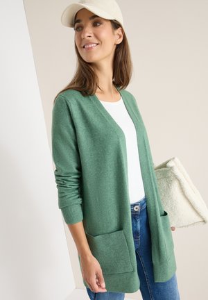 Groene cardigan met een relaxed fit, open voorkant en geribbelde mouwen. Voorzien van twee voorkruiken. Gecombineerd met een wit topje en een blauwe spijkerbroek.