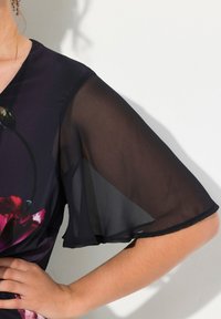 Černá květinová blůza s průsvitnými volánovými rukávy, se v-výstřihem a hladkým povrchem zdobeným barevnými květinovými vzory.