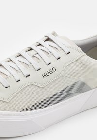 Vit nät-sneaker med vita snören, vit sula, svart nätaccent och "HUGO"-logotyp på sidan.
