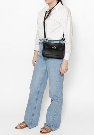 Borsa a tracolla in pelle nera con pattina e chiusura dorata, presentata su una persona che indossa una camicia bianca e jeans azzurri.