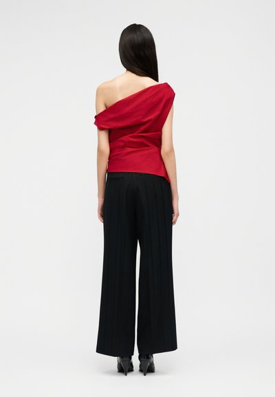 Γυναίκα με μακριά σκούρα μαλλιά που φοράει κόκκινο τοπ με ακάλυπτους ώμους και μαύρο παντελόνι φαρδιάς γραμμής, στέκεται γυρισμένη με την πλάτη σε απλό φόντο.