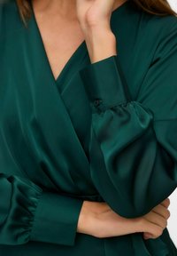 Zelena satenasta bluzica s pričvršćenim dizajnom, dugim napuhanim rukavima i dugmetima na manžetama, s glatkijom i sjajnom teksturom.
