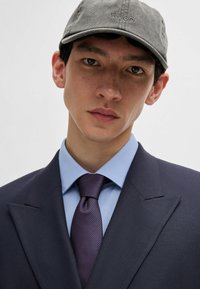 Dark blue suit jacket, light blue shirt, purple tie with a textured pattern, and a gray cap with "HUGO" embroidered on the front.