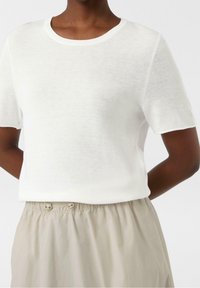 Mujer vistiendo una blusa de punto blanca de manga corta, sin estampados, metida dentro de unos pantalones elásticos de talle alto en color beige, con detalles de botones.