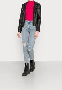 Person som bär en svart läderjacka, en ljusrosa tröja, slitna ljusblå jeans och svarta snörskor, står på en grå golv.