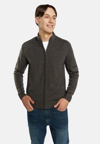 Donkergrijze zip-up trui met een ribbelstructuur, hoge kraag en lange mouwen, gecombineerd met blauwe spijkerbroek. Voorkant zakken zichtbaar.