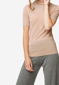 Beżowy sweter z krótkim rękawem typu golf wytworzony z miękkiego materiału. Tekstura jest gładka, a krój dopasowany. W zestawie z jasnoszarymi, luźnymi spodniami.