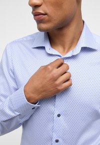 Hellblaues, gemustertes Hemd mit Button-Down-Kragen, strukturiertem Stoff und kontrastierenden schwarzen Knöpfen; Hand passt den Kragen an.