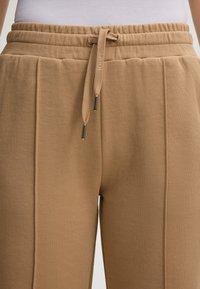 Khakifarbene Jogginghose aus weichem Material, mit elastischem Bund und Kordelzug, Seitennähten und Metallakzenten an der Schnur.