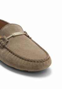 Suede mokkasiner i lysebrun med rund tå, syningdetaljer og en guldfarvet metalaccent over vamps. Blød tekstur.
