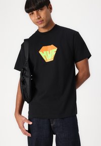 Zwart T-shirt met korte mouwen met een oranje en groene ruitlogo met de tekst "HUF," gecombineerd met een donkere spijkerbroek en een zwarte tas.