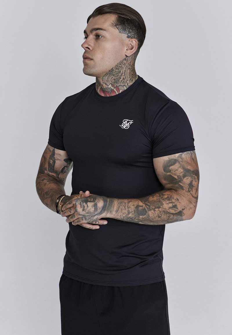 Melnas pieguļošas T-krekls, kas izgatavots no gludas, elastīgas auduma, īsām piedurknēm, ar mazu baltu logo uz krūtīm. Pozē modelis ar tetovējumiem.